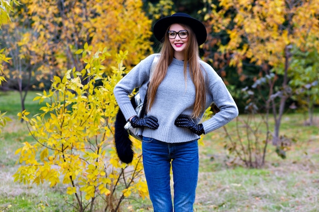 Good-looking woman with long hair wears jeans and gloves standing in confident pose on nature background. Outdoor photo of pretty female model in trendy gray sweater walking in park in autumn day.