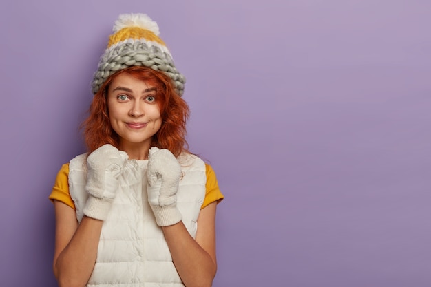 Free photo good looking young female raises clenched fists, wears vest, t shirt, white gloves, headgear on head, looks with intriguing gaze at camera, isolated on purple wall