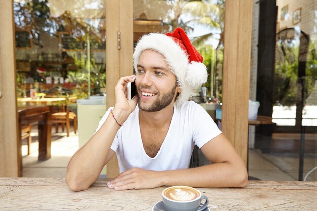 Good-looking young man with stubble having cappuccino sitting at wooden cafe table and greeting friends on Christmas, using smart phone