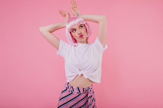 Free Photo good-looking young woman in casual stylish t-shirt fooling around . indoor portrait of romantic lady in pink peruke making faces on bright wall