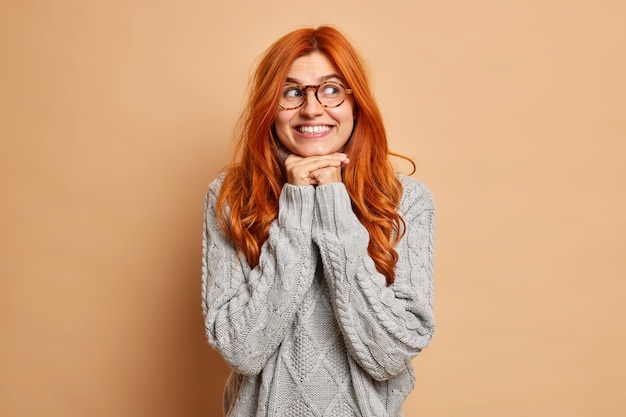 Good looking young woman with red hair keeps hands under chin smiles toothily concentrated aside wears spectacles and sweater.