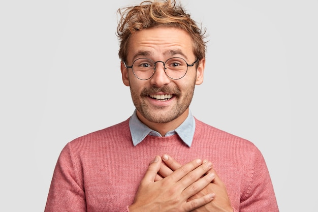 Free photo good natured hipster man keeps hands on chest, has positive facial expression, curly trendy hairstyle, being thankful to guests