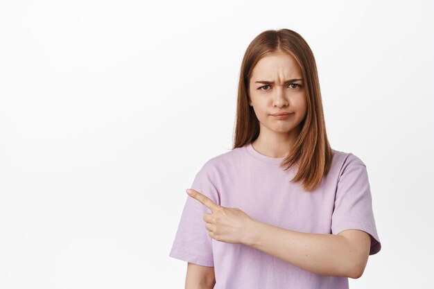 Not good. Portrait of blond girl furrow eyebrows from dislike, pointing finger left and complaining, disappointed by smth bad, condemn, standing against white background.