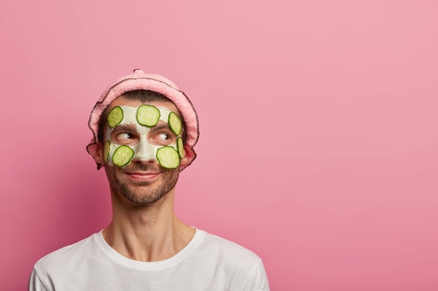 Free photo good tempered happy man wears face mask and cucumbers, enjoys morning spa procedure, wants to look refreshed