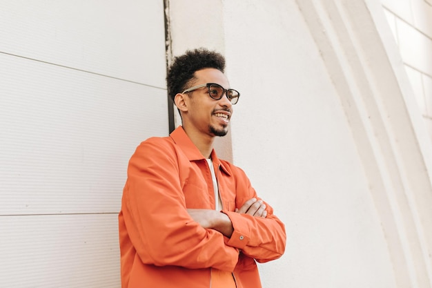 Free photo goodhumored curly bearded darkskinned man in stylish orange jacket and sunglasses smiles crosses arms and leans on white wall outside