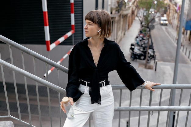 Free Photo goodhumored woman standing outside shorthaired girl in black shirt and white pants in wireless headphones holding a bottle of water and smiles outdoors