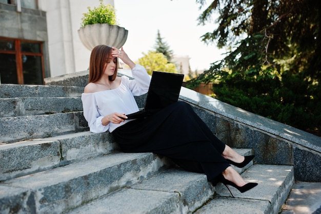 Goodlooking young woman in white blouse wide black pants and black classic high heels sitting on stairs and working on her laptop