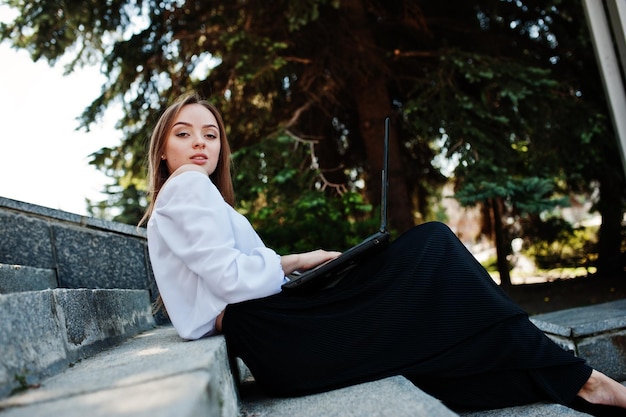 Goodlooking young woman in white blouse wide black pants and black classic high heels sitting on stairs and working on her laptop