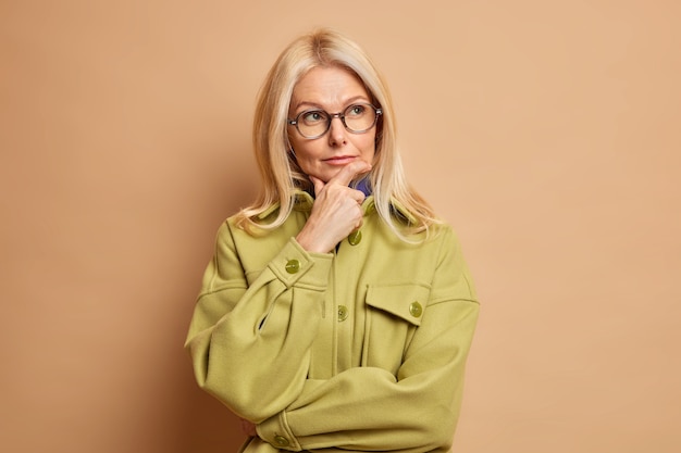 Free photo gorgeous attractive woman of middle age holds chin and thinks deeply turns gaze away dressed in fashionable jacket ponders on something important.