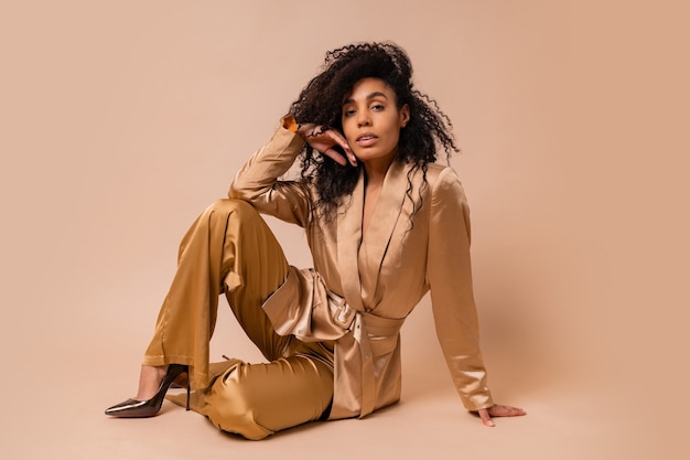 Gorgeous  black woman with beautiful wavy hairs in elegant  golden satin suit posing over beige wall. Spring fashion look.