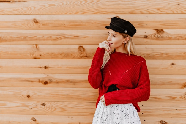 Gorgeous blonde woman in black hat and nice red sweater posing tenderly on the wooden wall. Charming girl enjoying warm autumn day outside.