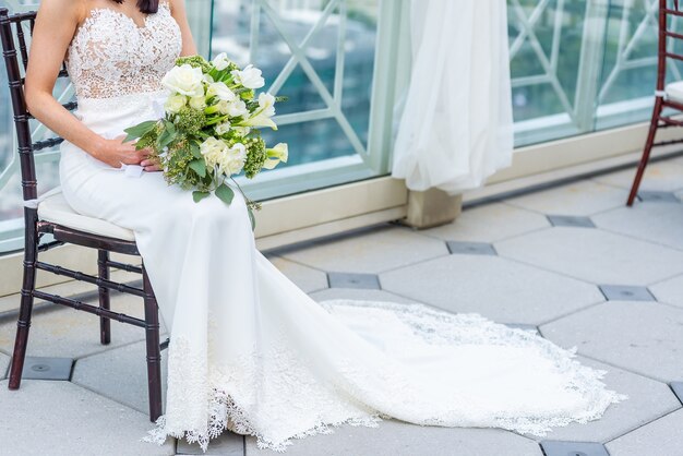 Gorgeous bride with a luxury wedding dress sitting on a chair holding a bridal bouquet