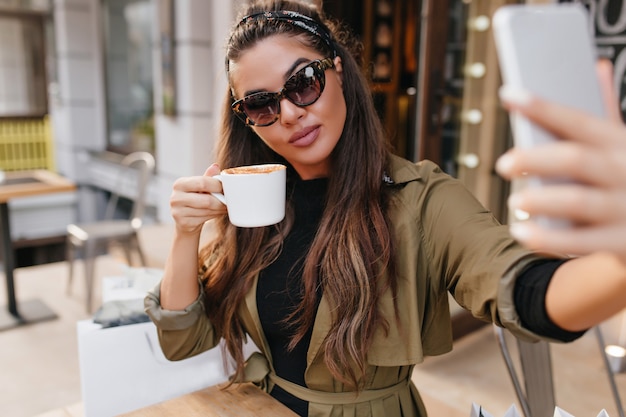 Free photo gorgeous brunette woman in sunglasses drinking coffee and taking picture of herself in weekend