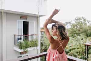 Free photo gorgeous caucasian girl in pink attire stretching at hotel balcony. magnificent curly woman enjoying city view from terrace.