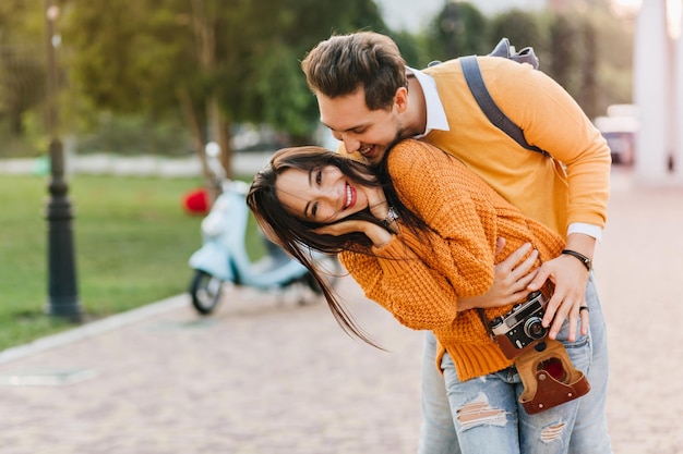 Free photo gorgeous female model in ripped blue jeans fooling around with husband after photoshoot in autumn park. blissful girl in knitted sweater with camera having fun with boyfriend enjoying weekend together