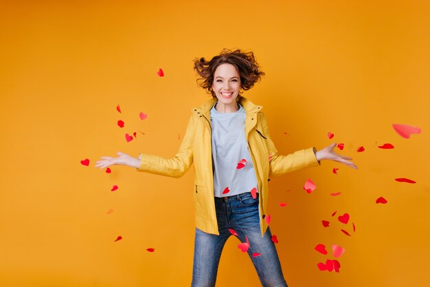 Free photo gorgeous female model throwing out paper hearts and expressing happiness. glamorous curly woman celebrating valentine's day.