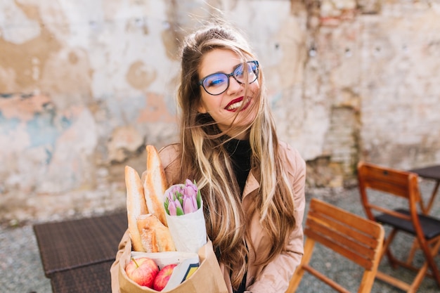 Gorgeous girl after photosession bought fresh food and drinks coffee enjoying a sunny day. Stylish young female photographer holding grocery bag and cup of cappuccino posing in the outdoor cafe.