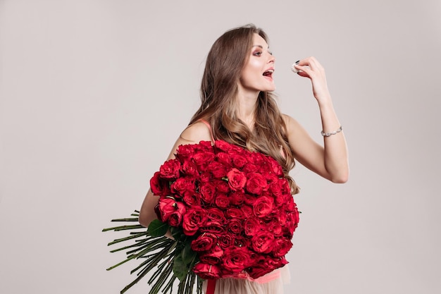 Free photo gorgeous happy brunette with long wavy hair holding beautiful red roses bunch and eating delicious dessert with her hand isolated on white background in studio