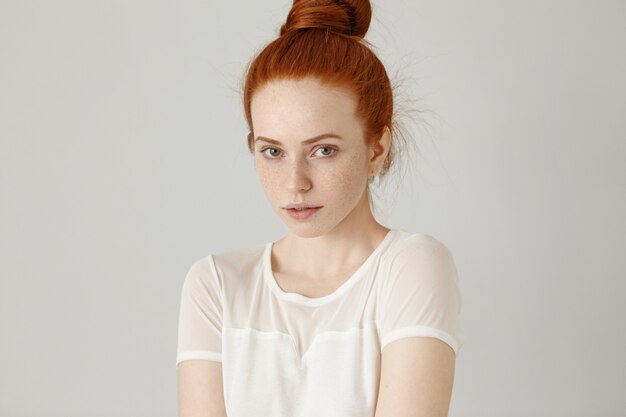 Gorgeous redhead girl with hair knot and freckles dressed in white blouse, shrugging her shoulders slightly with uncertainty, looking, having cute shy smile