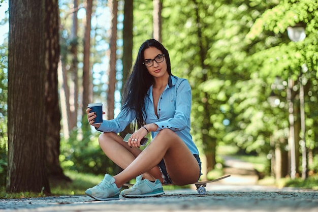 Free photo gorgeous sexy brunette girl in glasses wearing a shirt and shorts sitting on a skateboard and holds cup of takeaway coffee and smartphone at a park.