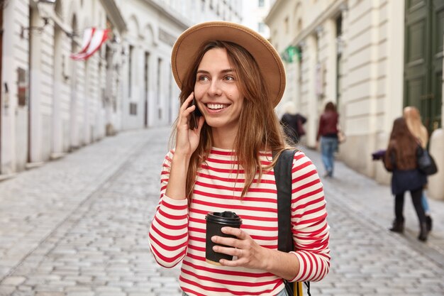 Gorgeous smiling tourist strolls around city, has phone conversation, holds takeaway coffee, focused somewhere into distance