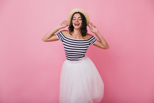 Gorgeous tanned female model posing in trendy lush skirt and striped t-shirt. Indoor portrait of refined latin girl touching her hat.