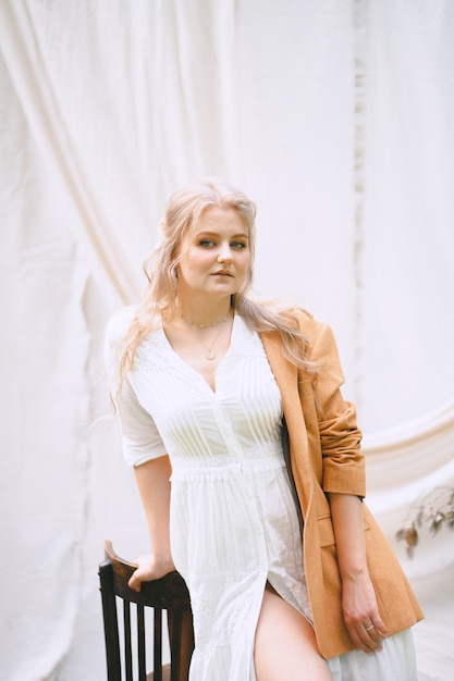 Gorgeous woman standing and looking in garden with white wall in white dress and brown jacket during daytime .