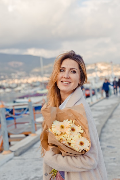 Free photo gorgeous woman standing and smiling in seaside with flowers during daytime and and looking lovingly.