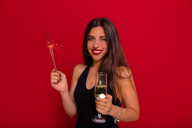 gorgeous woman with dark long hair wearing black dress with bare shoulders holding a glass of champagne and sparkler preparing for Christmas party over red wall