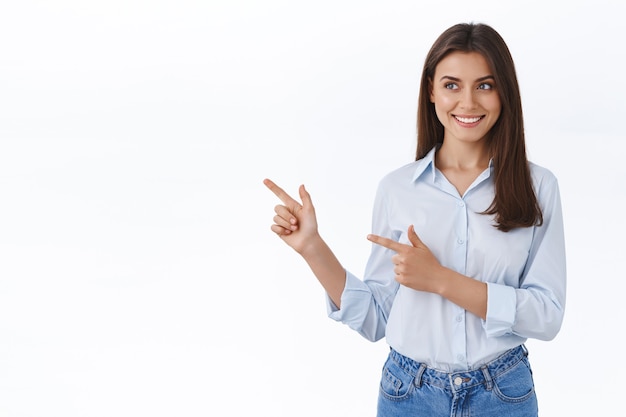 Gorgeous young adult woman in blue blouse introduce her company banner, friendly and happy smiling, pointing looking left at blank white space for your advertisement, standing studio wall