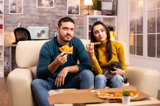 Free photo gorgeous young couple eating pizza while watching tv in the living room sitting on the sofa