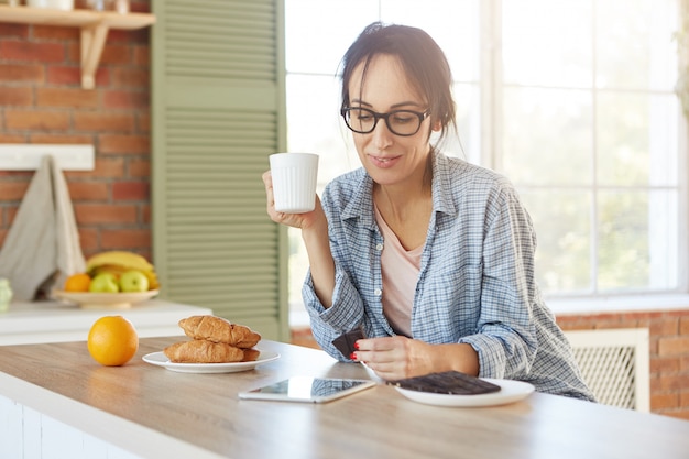 Gorgeous young female model wears spectacles and shirt, drinks coffee with croissants and dark chocolate, has breakfast before work,