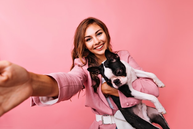 Free photo gorgeous young woman in pink jacket taking picture of herself with dog. lovely caucasian girl playing with bulldog.