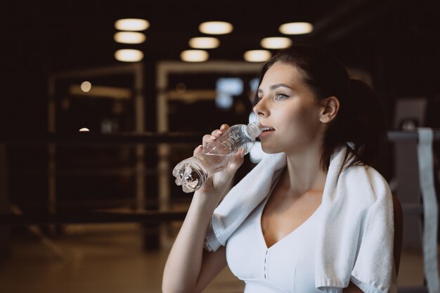 Gorgeous young woman with a towel on her shoulders drinking water from a bottle at the gym