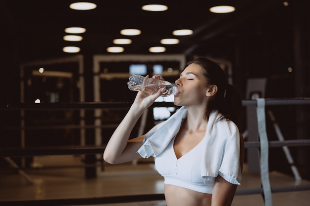 Gorgeous young woman with a towel on her shoulders drinking water from a bottle at the gym