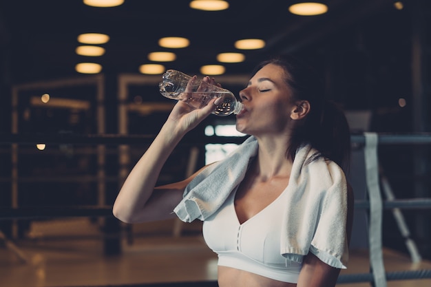 Gorgeous young woman with a towel on her shoulders drinking water from a bottle at the gym