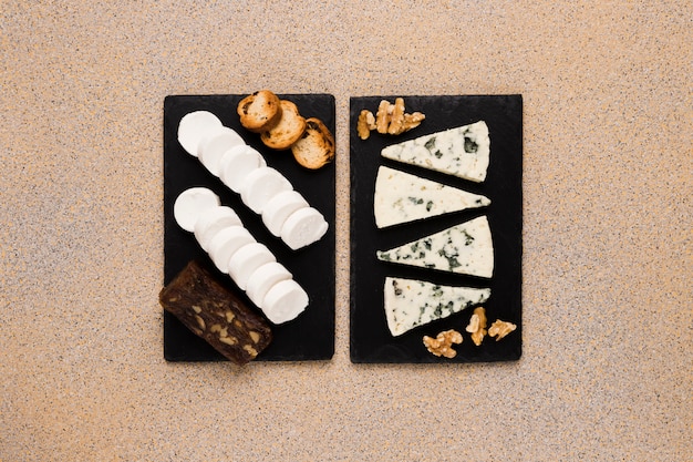 Free photo gorgonzola cheese slices and walnut with goat cheese; brown cheese and bread on black slate stone over textured wallpaper