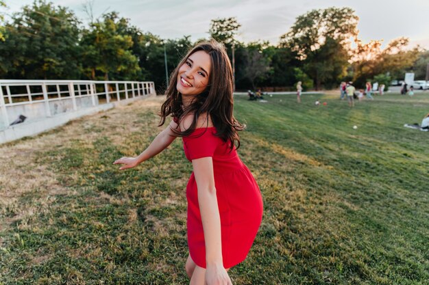 Graceful dark-haired girl expressing happiness . Outdoor shot of gorgeous brunette woman in red dress having fun in park.