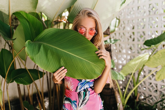 Graceful fair-haired girl wears elegant sunglasses hiding behind big leaf.