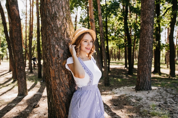 Graceful girl in straw hat enjoying good day in summer. Outdoor photo of refined female model posing with smile in forest.