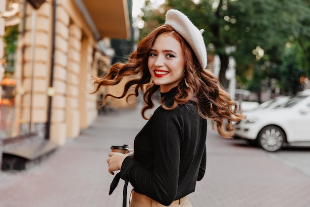 Graceful long-haired ginger girl looking over shoulder. Laughing pretty woman in beret enjoying walk.