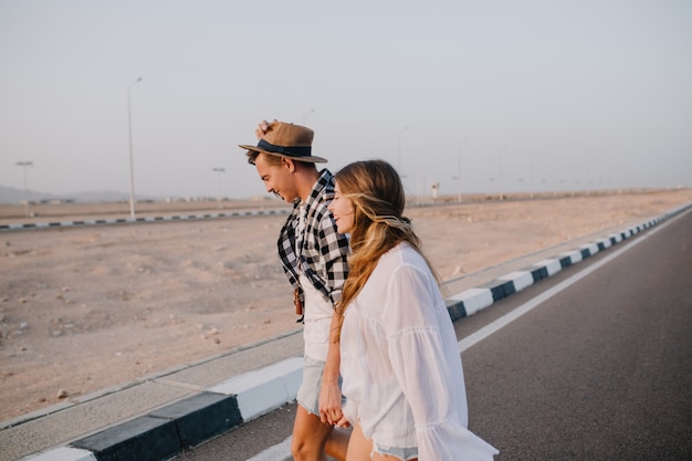 Graceful long-haired woman in white shirt and boy in hat walking down the highway holding hands and smiling. Stylish couple crosses the road and talking about travel under open sky in early morning