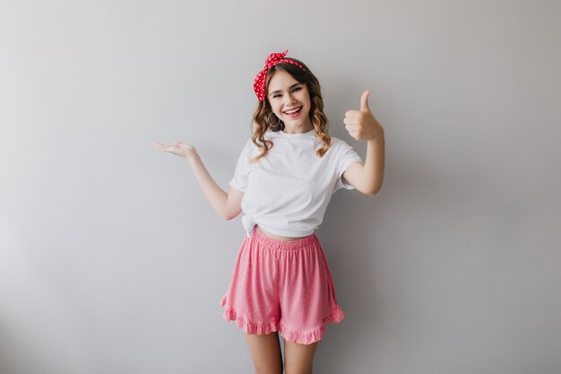 Graceful positive girl in pajamas posing with thumb up. Indoor shot of enthusiastic young lady expressing happiness