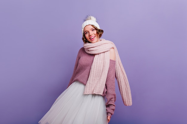 Free photo graceful white girl in long warm scarf standing on purple wall.   refined pleased young woman in hat posing  with smile.