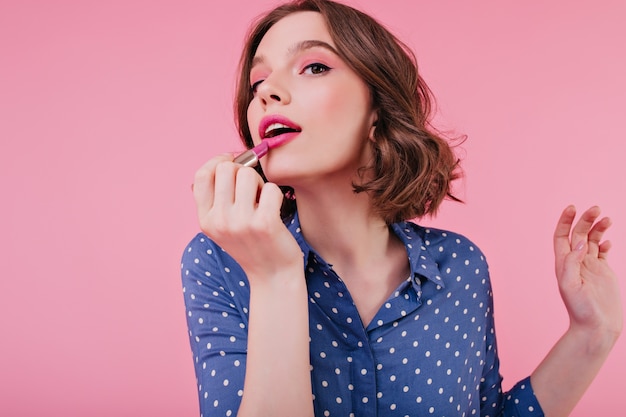 Free photo graceful woman doing lips makeup on bright wall. indoor portrait of magnificent short-haired girl posing with lipstick.