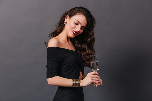 Graceful woman with wavy long hair and in stylish black dress posing with champagne on dark background.