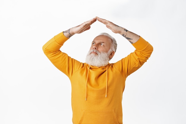 Free photo grandfather with tattoos looking confused at his hands roof gesture, frowning thoughtful while making home rooftop, standing against white wall