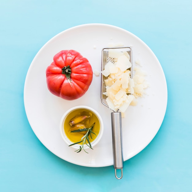 Free Photo grated cheese with whole red tomato and oil on white plate over the blue background