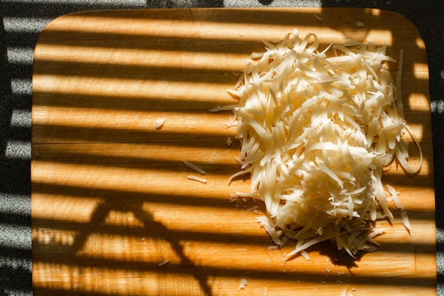 Grated cheese on wooden board. Stripes from blinds on the window casting over board with shredded cheese.