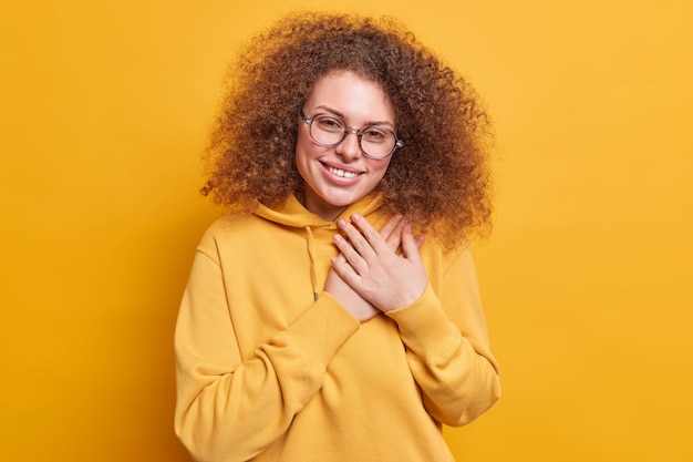 Free photo grateful pleased curly haired young woman holds hands near heart smiles pleasantly says thank you appreciates help dressed in sweatshirt isolated over yellow wall. body language concept